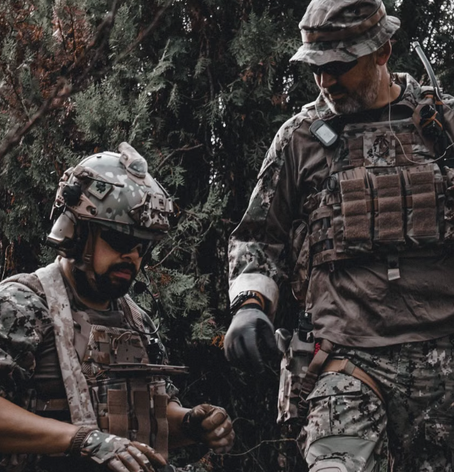 Two tactical operators in camouflage gear coordinating during a military field exercise in a wooded environment